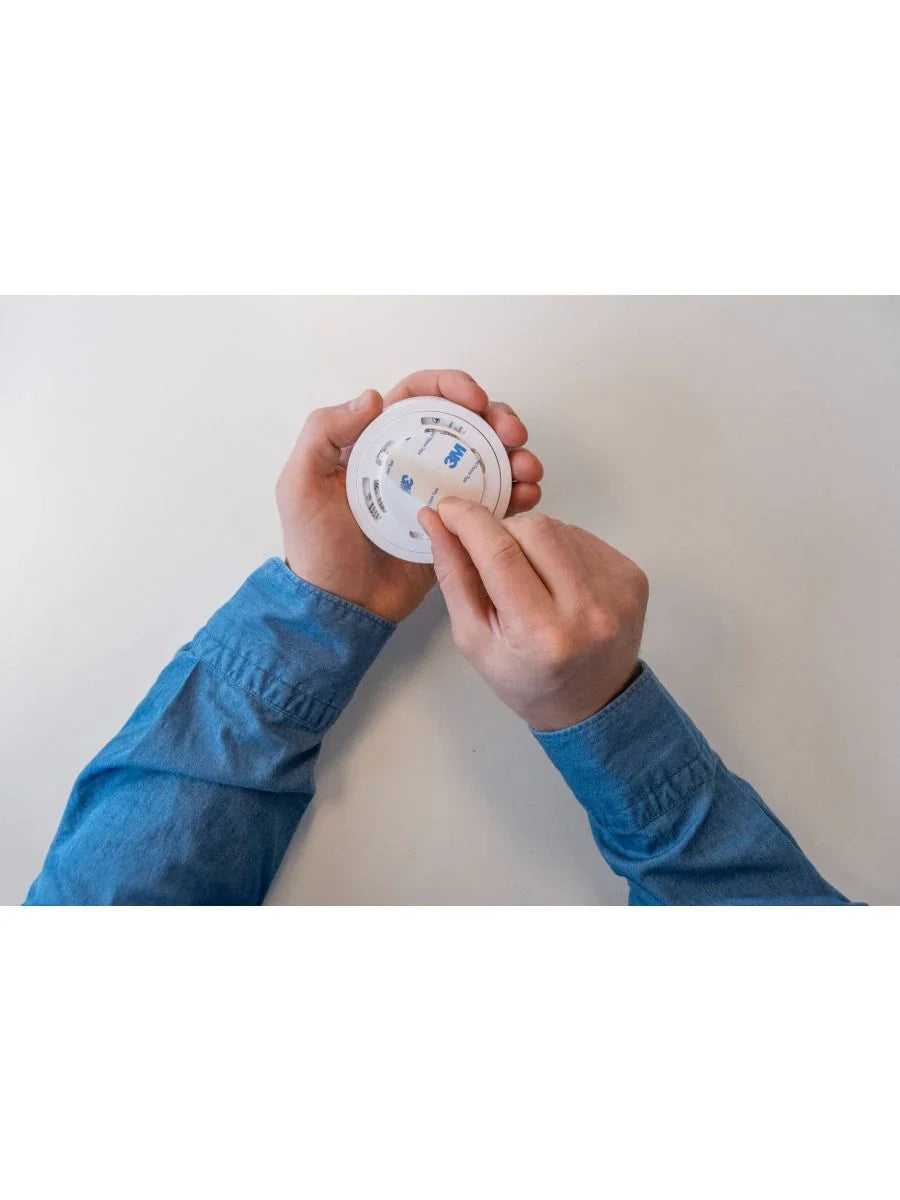 Person installerer rund røykvarsler på hvit vegg med selvklebende tape, brannvernutstyr