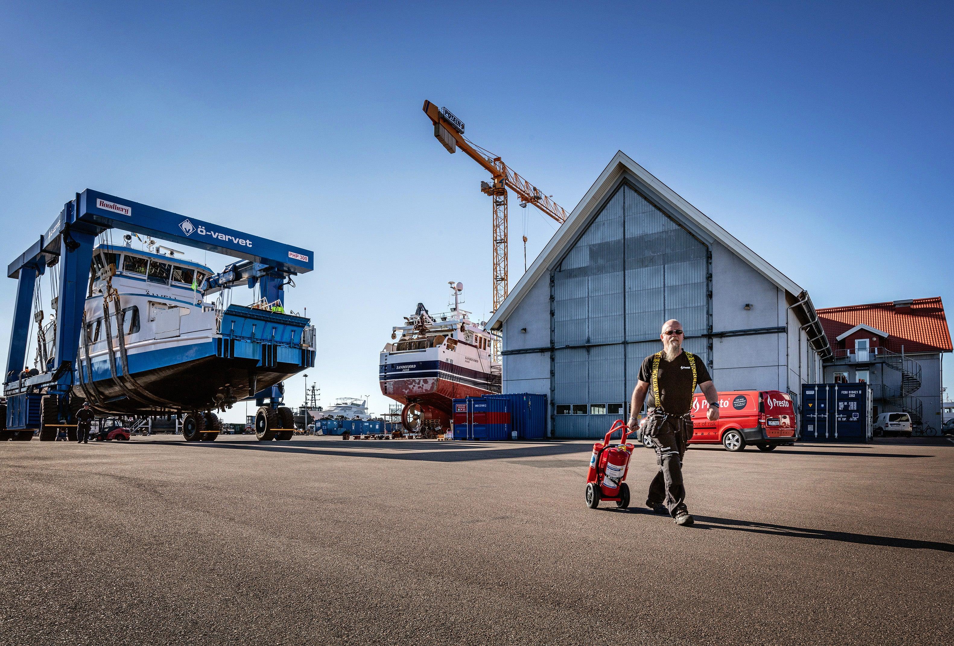 Mann triller brannslukker på havneområde med båter, industri, brannsikkerhetsutstyr