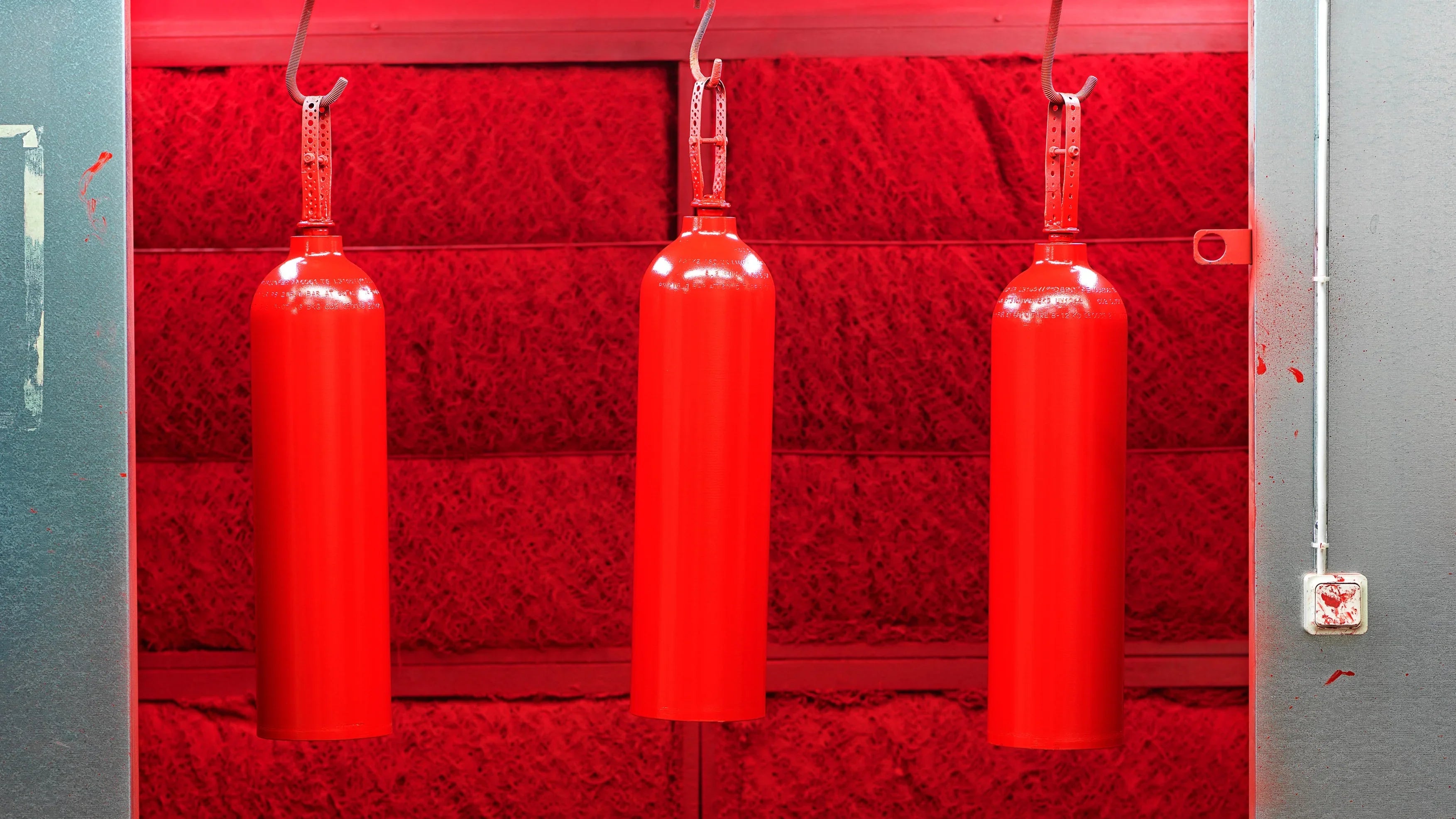 Tre brannslukkere henger til tørk etter lakkering, industrielt verkstedmiljø.