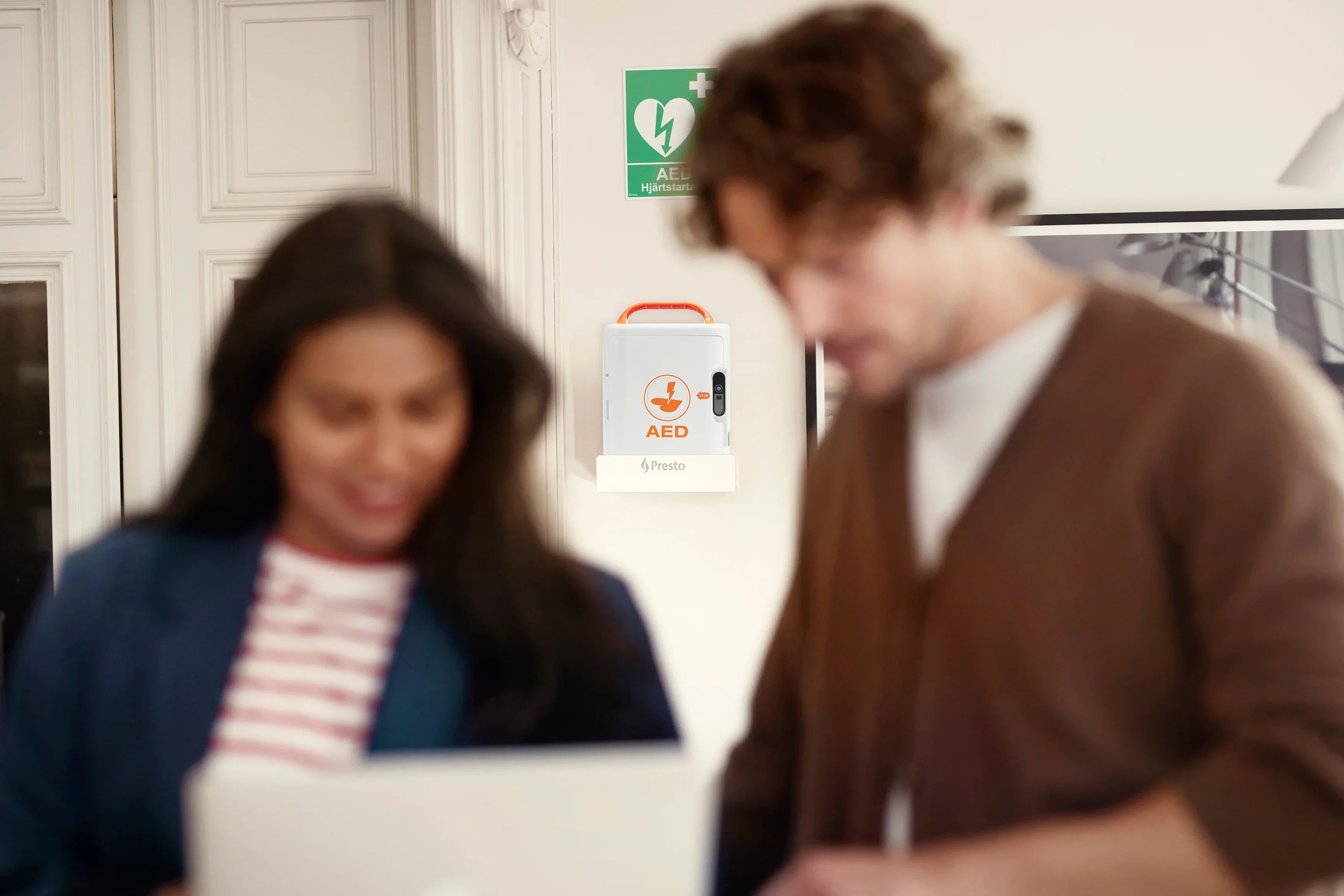 Vegghengt hjertestarter i kontormiljø med to personer foran, fokus på førstehjelp